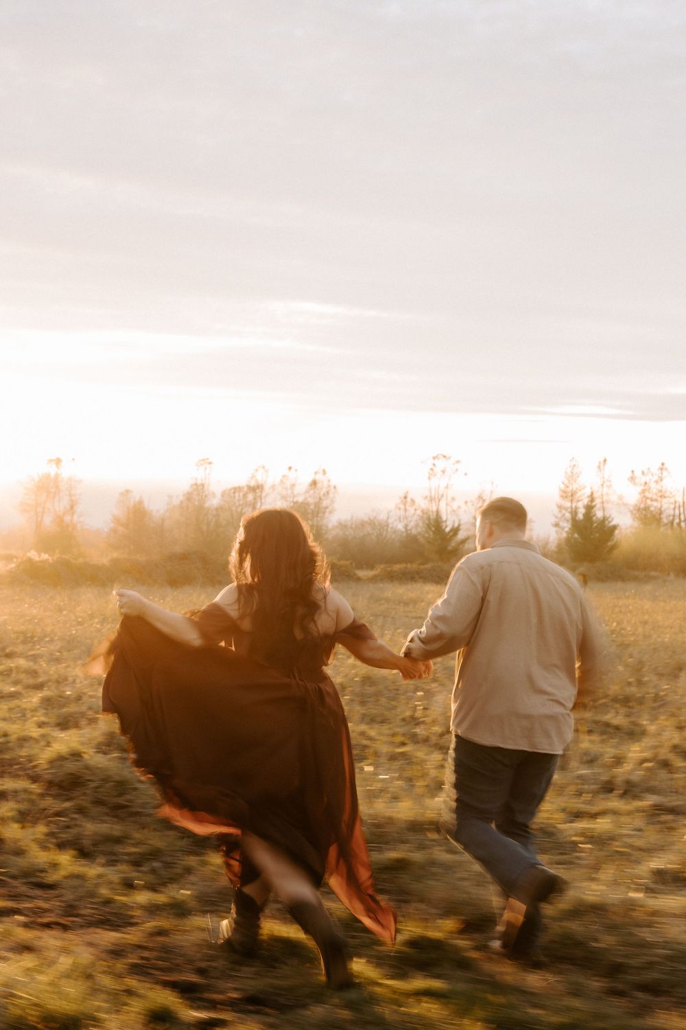 A couple runs together through a golden field at sunset as her flowing dress catches the warm evening breeze.