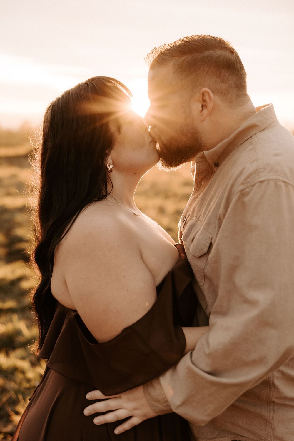 An intimate couple embraces in warm sunset light on a grassy hillside during a romantic photoshoot.