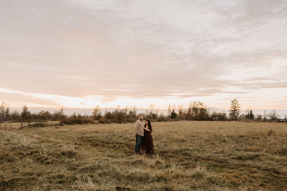 Two figures stand together in an open field at sunset with dramatic cloudy skies and trees in the background.