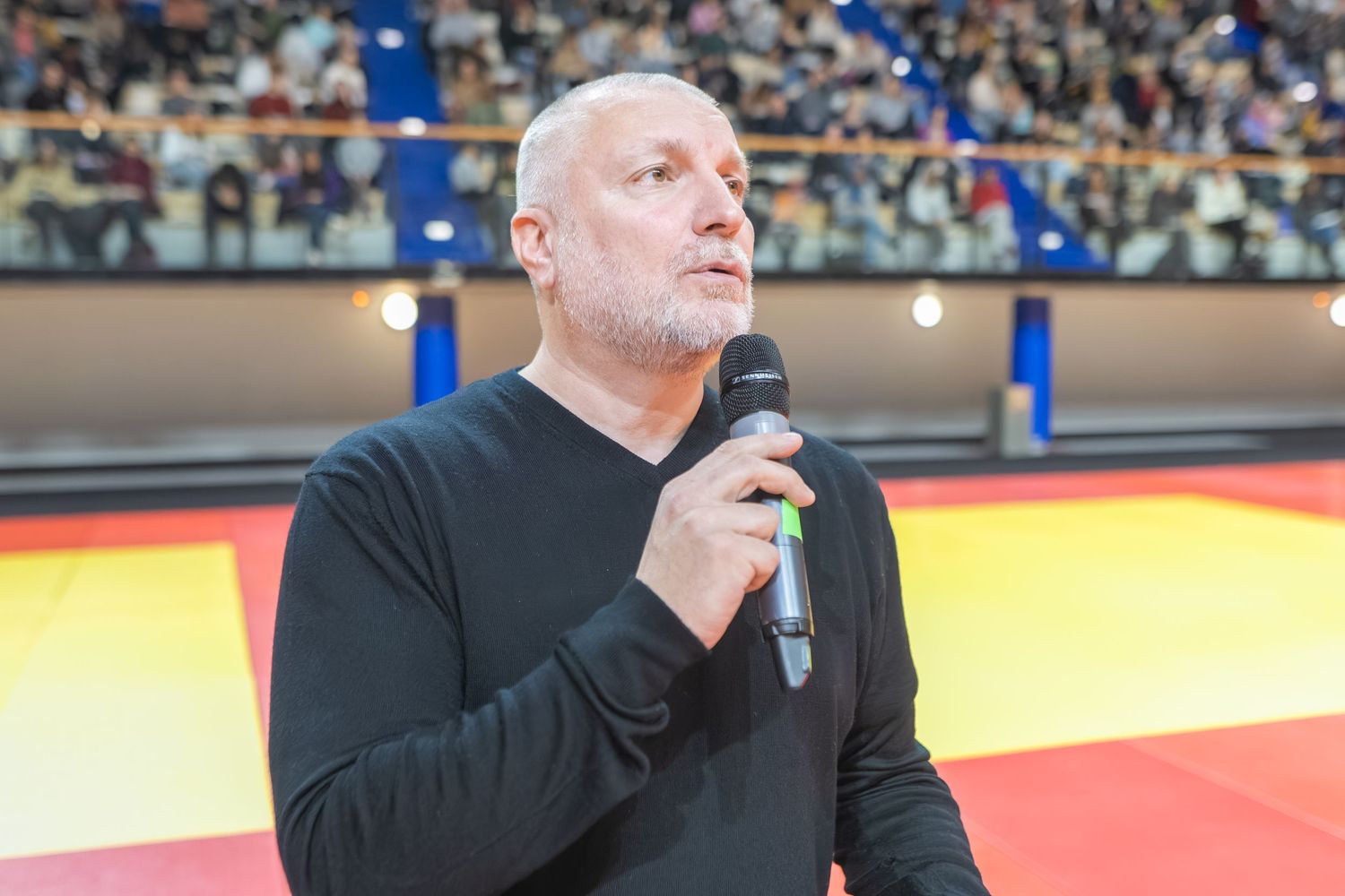 A person in a black jacket speaks into a microphone at a sports event with red and yellow lighting visible.