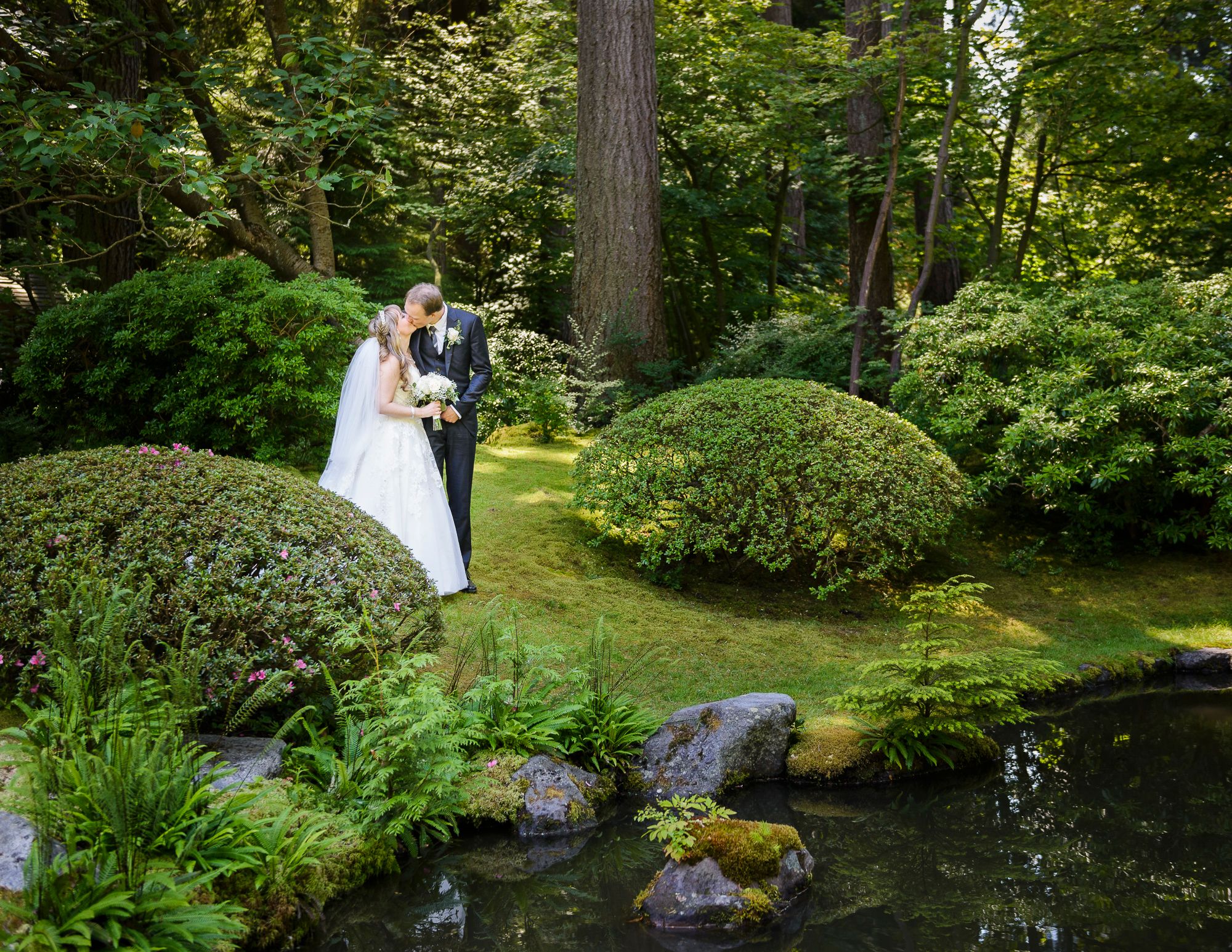 A peaceful garden path leads through lush greenery and over a small wooden bridge near a tranquil pond.