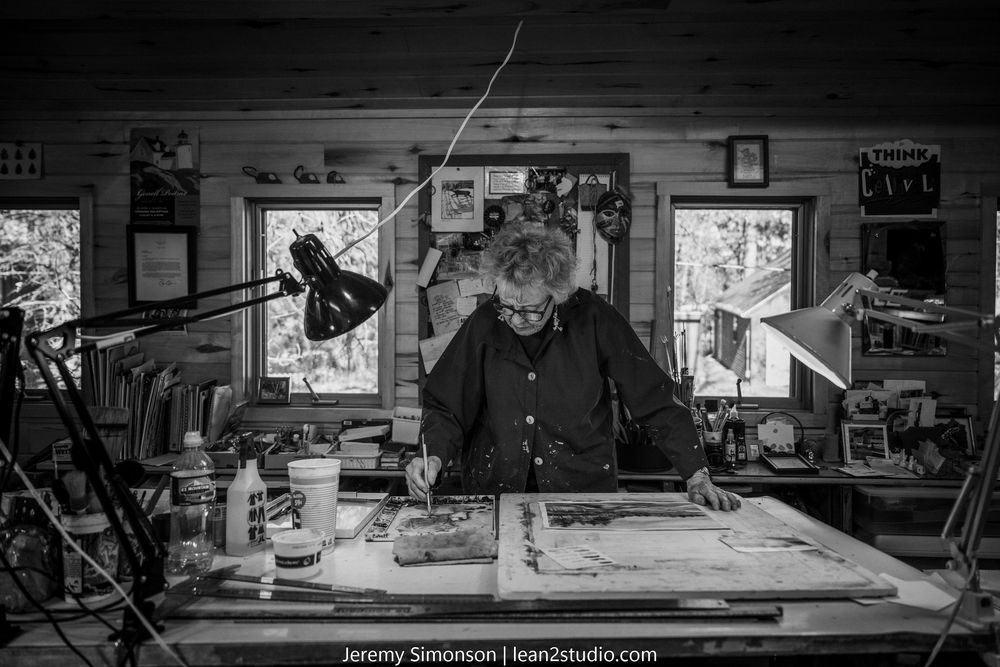 A person works at a cluttered desk in a dimly lit workshop with light streaming through windows in black and white.