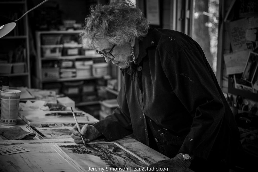 A black and white photo shows a person working at a desk with papers and documents spread across it.