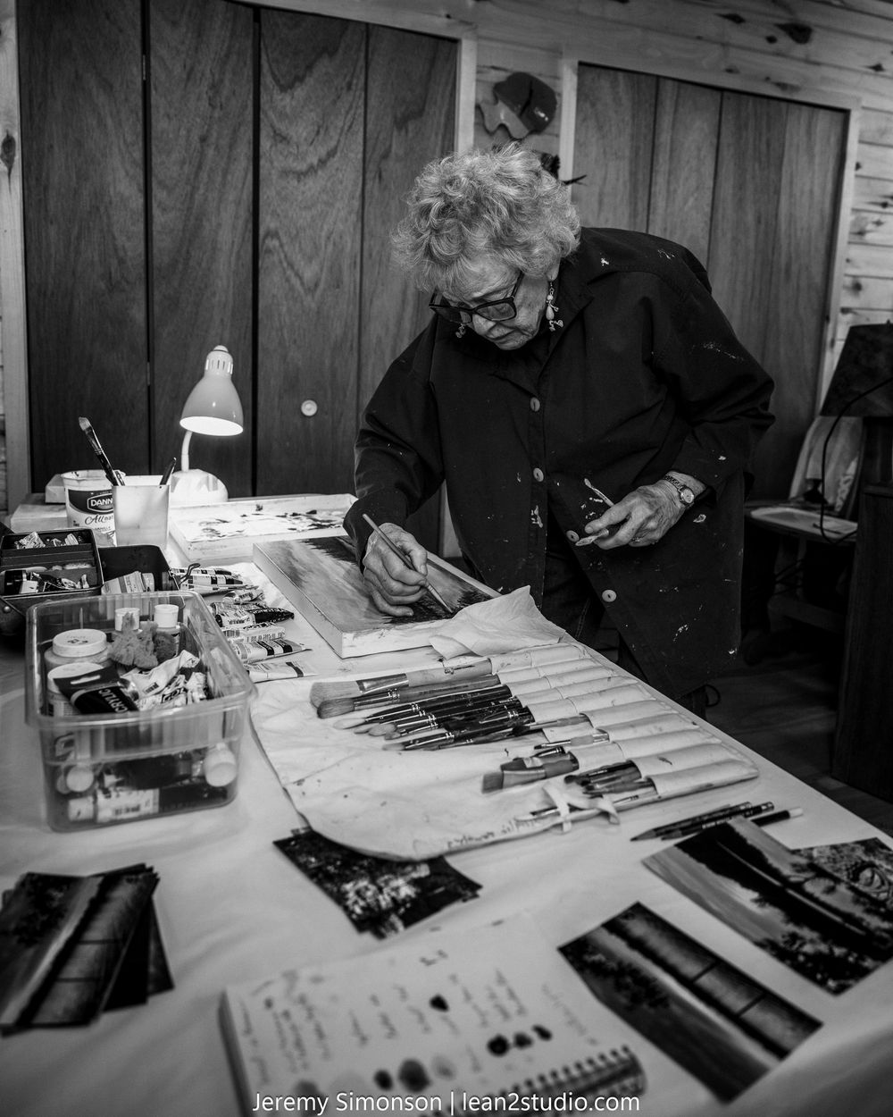 Black and white image showing someone working at a table with art supplies and tools spread out in a studio space.