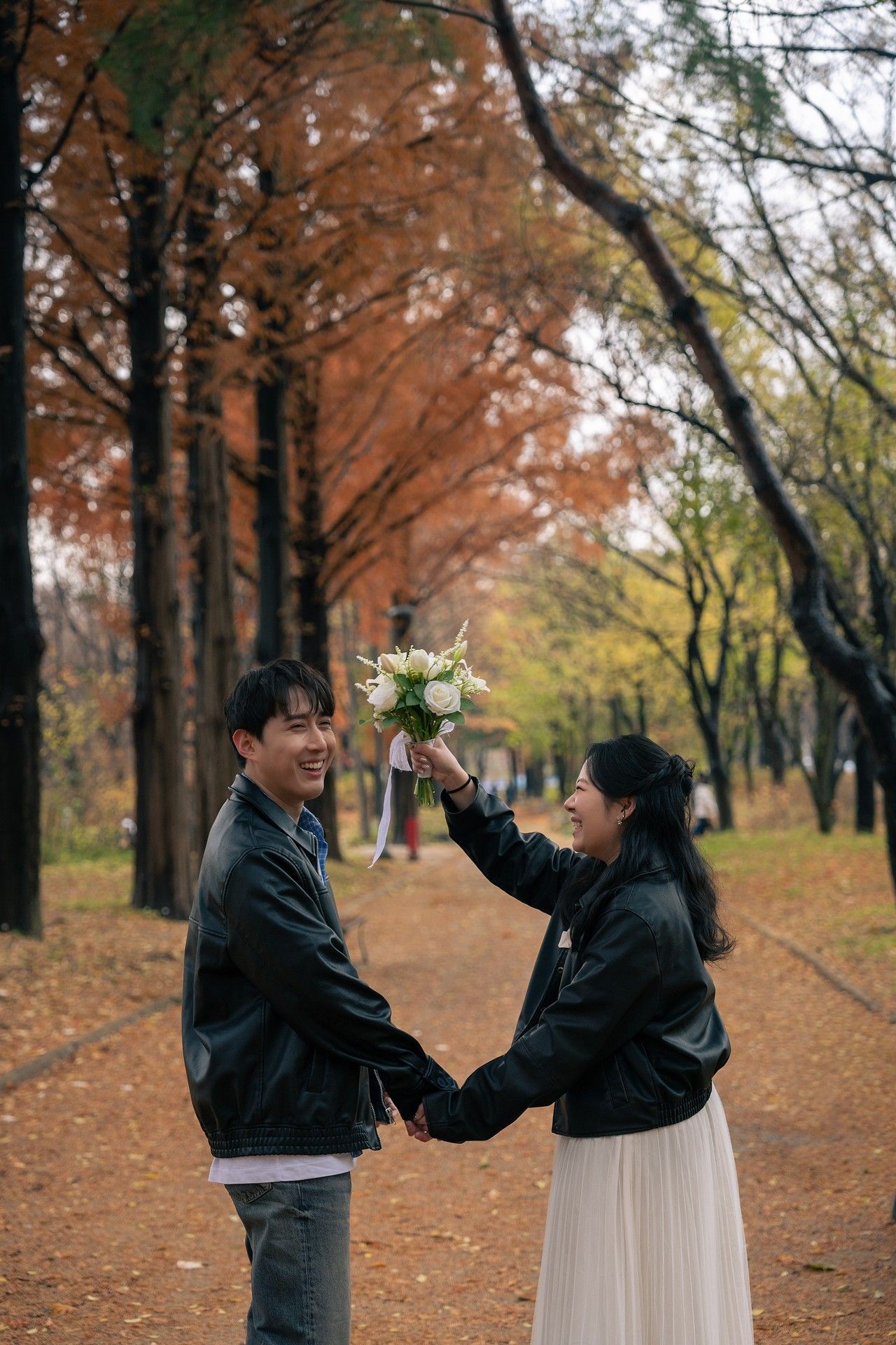 A couple in black leather jackets playfully interacts in a park with autumn foliage in the background.