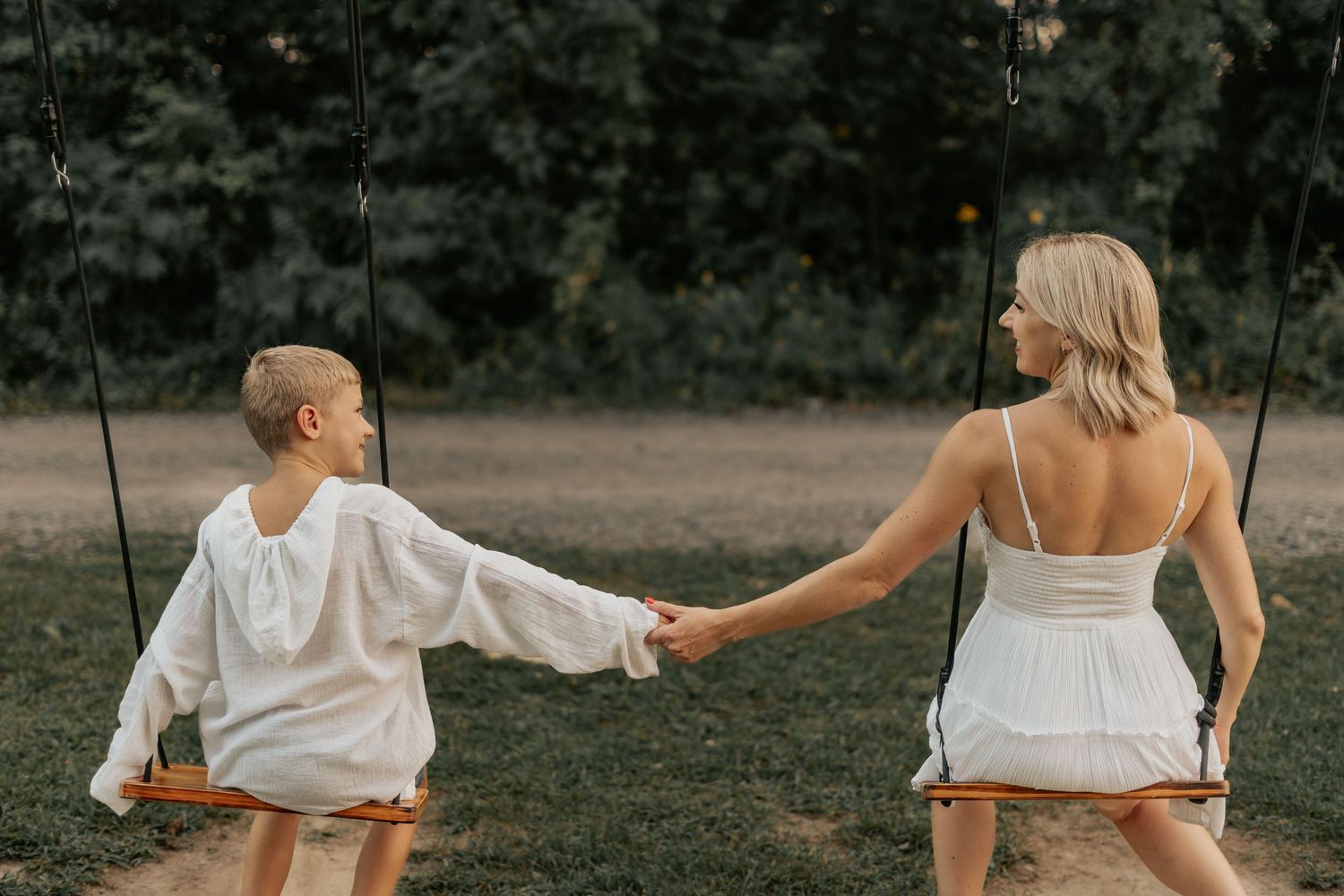 Two people in white summer outfits holding hands while sitting on swings in a grassy outdoor setting at dusk.