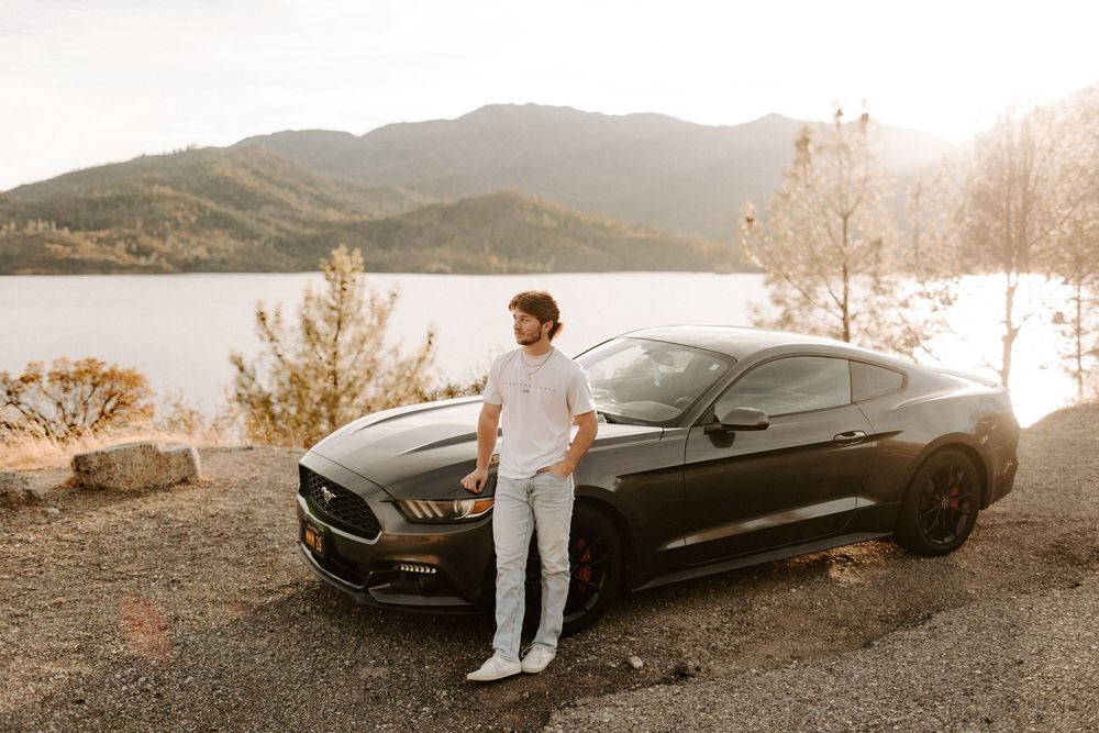 A sleek black sports car parked on a scenic mountain road during golden hour with a figure standing nearby.