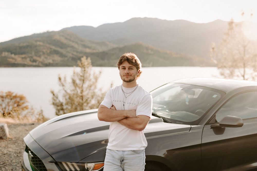 A person stands beside a sleek black sports car against a scenic mountain landscape at sunset.