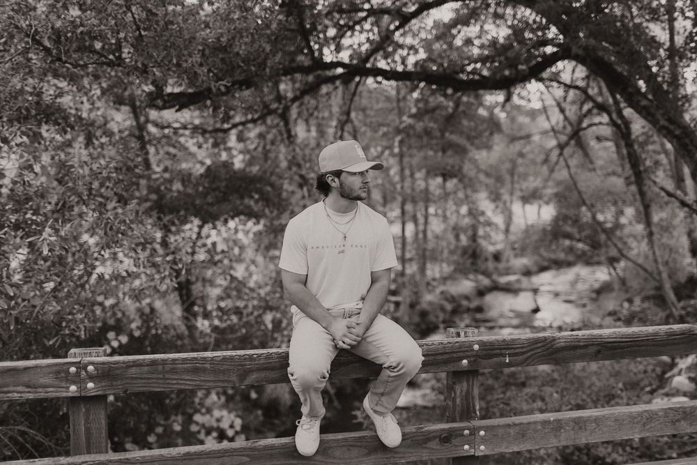 A person in casual attire sits on a wooden fence post in a wooded outdoor setting.