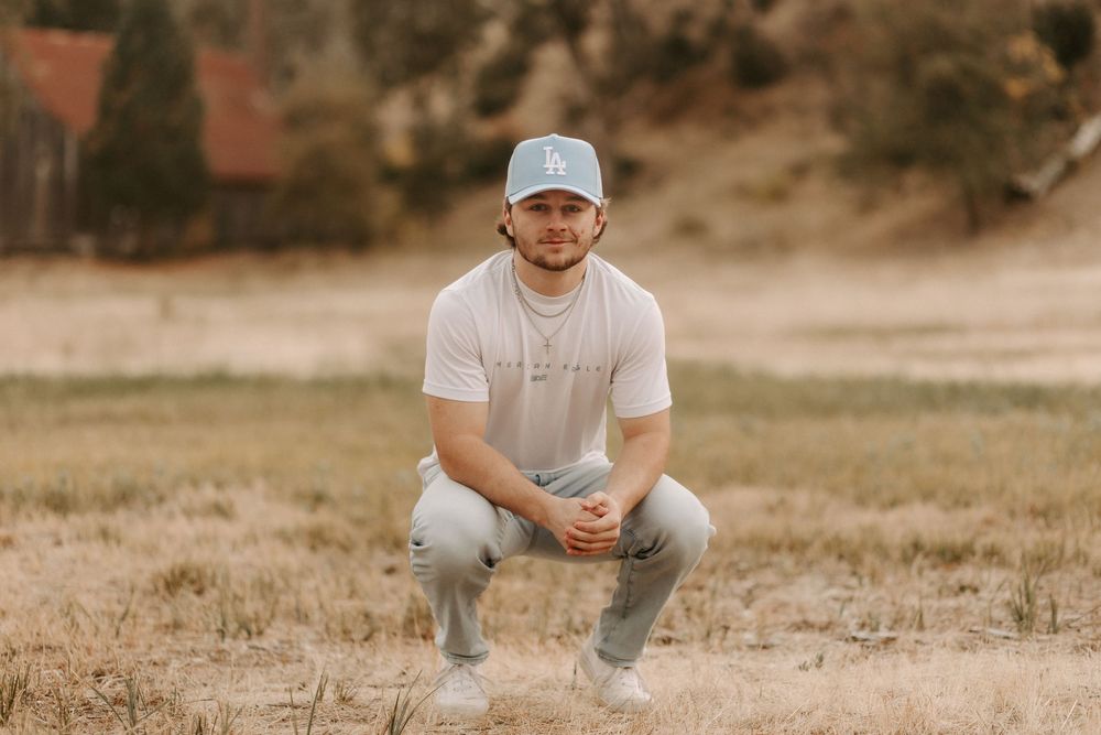 A person in a baseball cap and white t-shirt crouches in a grassy field during golden hour.