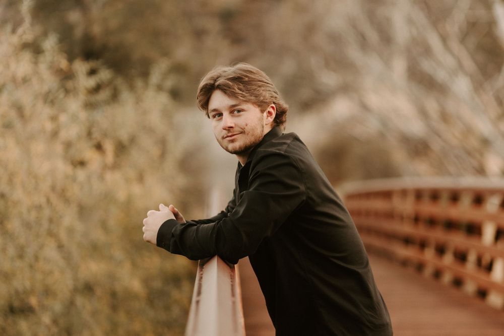 Person in black coat poses thoughtfully on wooden bridge in autumn setting.