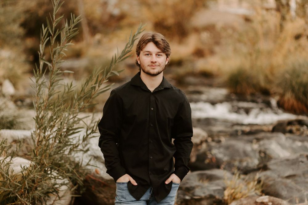A person in a black sweater sits thoughtfully among tall grasses and autumn foliage in a natural outdoor setting.