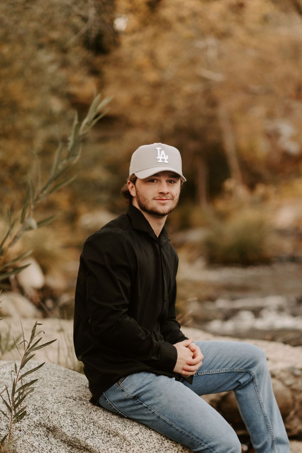 Person in LA baseball cap and black jacket sits casually on rocky terrain during autumn.