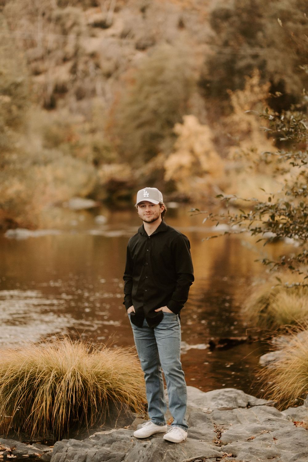 Person in casual attire poses on rocks by a serene river with golden autumn foliage in background.