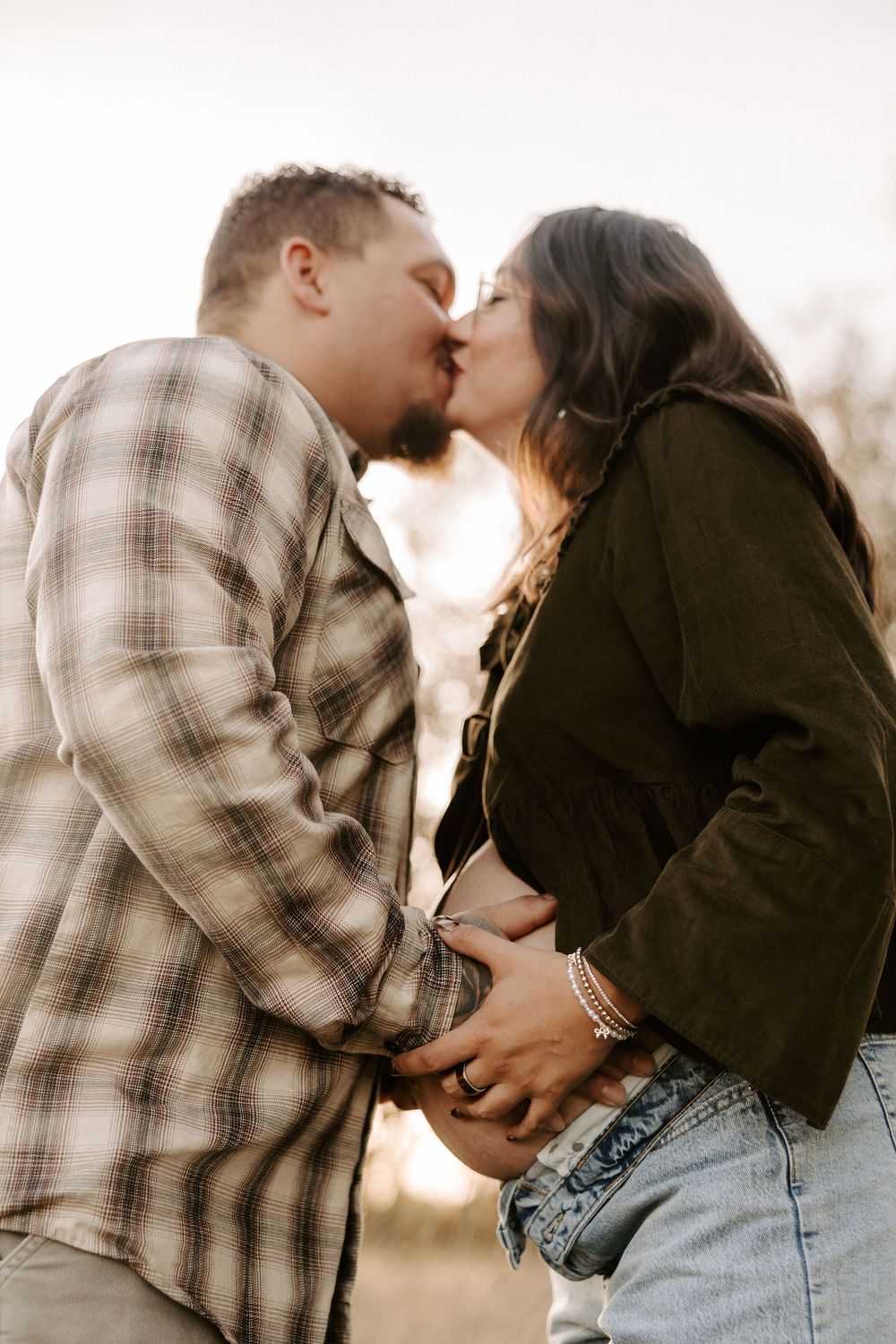 A couple sharing a romantic kiss outdoors at sunset, dressed casually in plaid and denim.