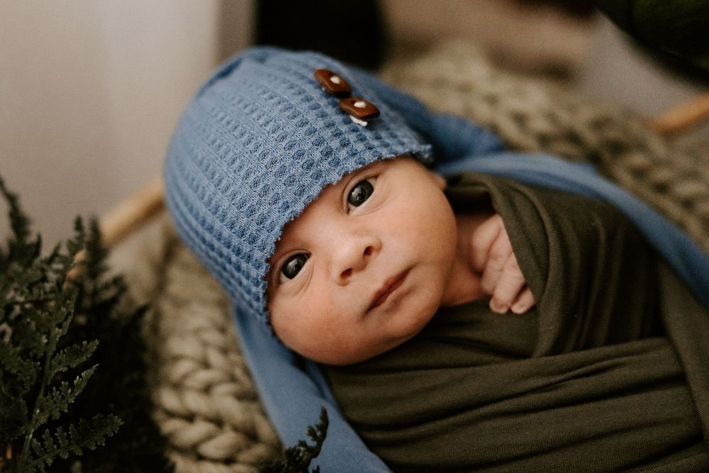 A baby wearing a blue knit hat and gray sweater rests peacefully on a beige knit blanket.
