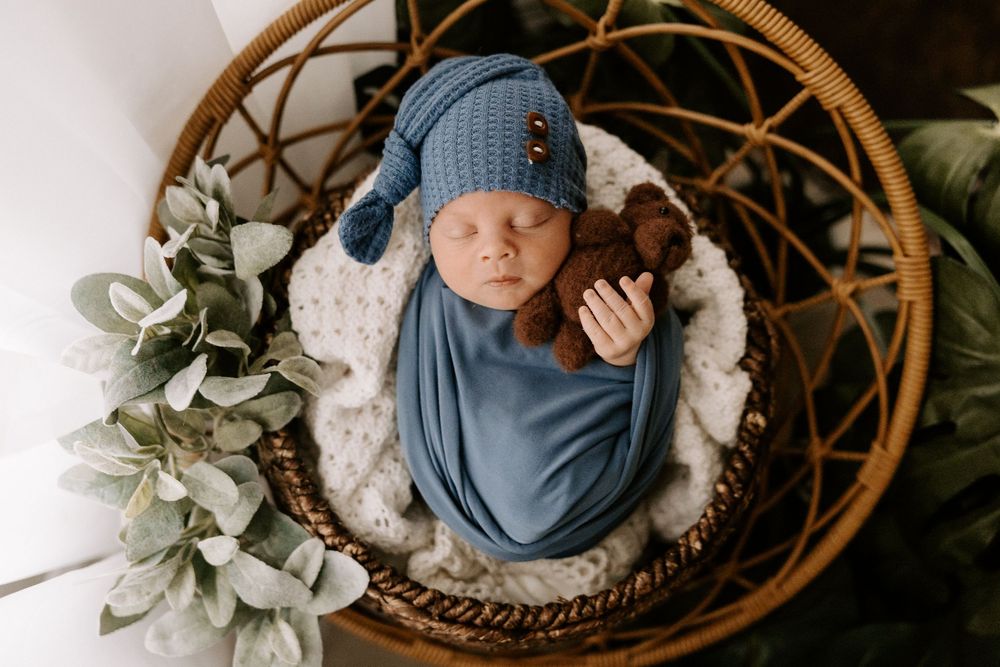 Newborn baby wrapped in blue swaddle blanket sleeping peacefully in woven basket with eucalyptus decor.