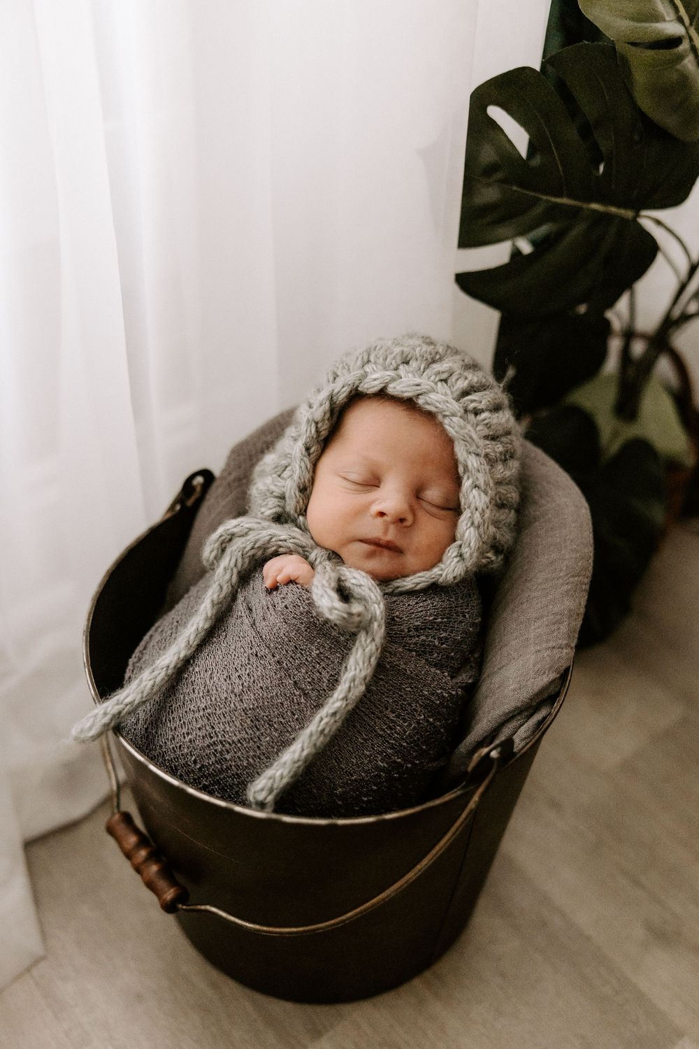 A sleeping newborn wrapped in a gray knit blanket and bonnet rests peacefully in a dark metal bucket near a houseplant.