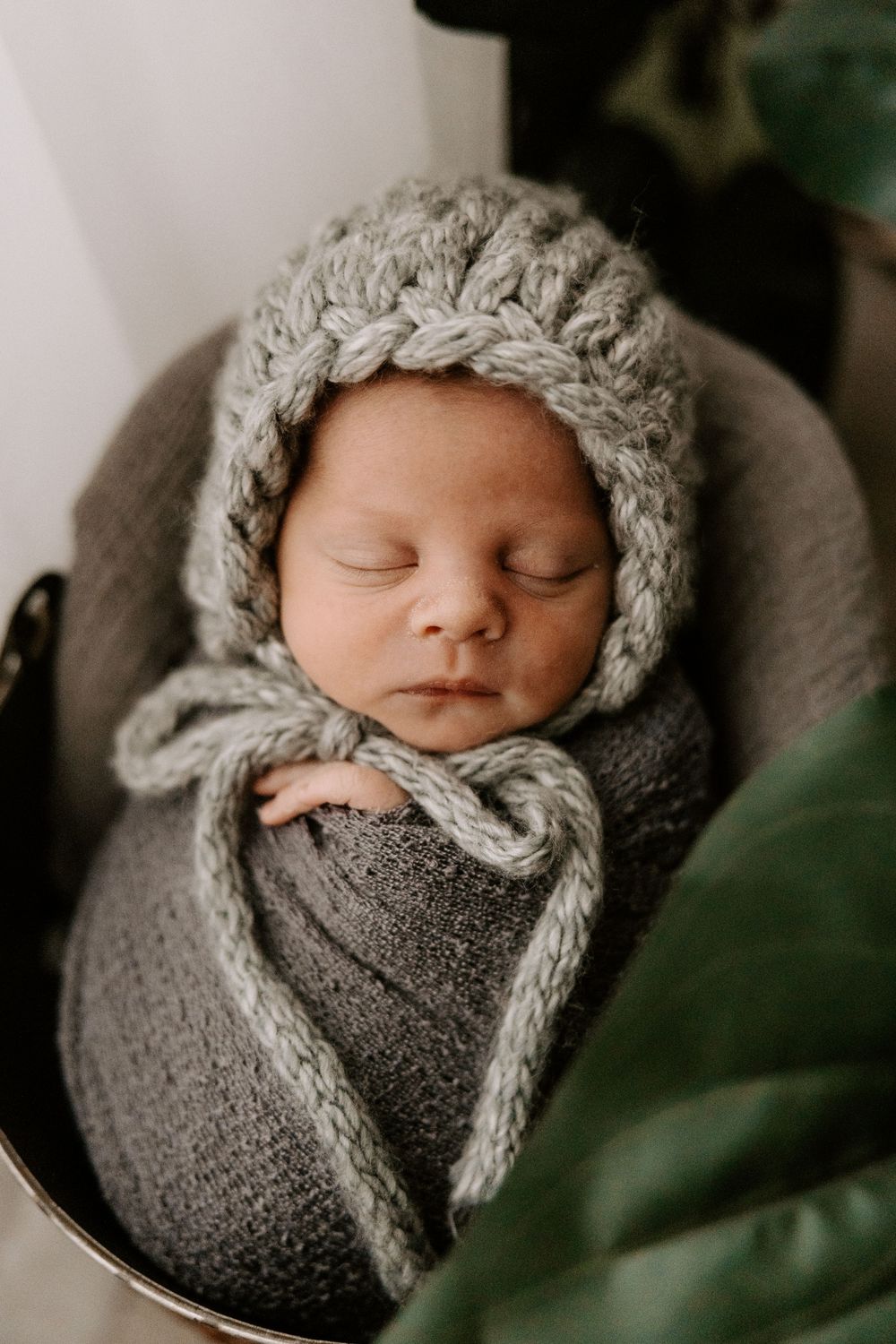 A sleeping newborn baby wrapped in gray knit swaddle and wearing a cozy winter bonnet in a professional photo series.