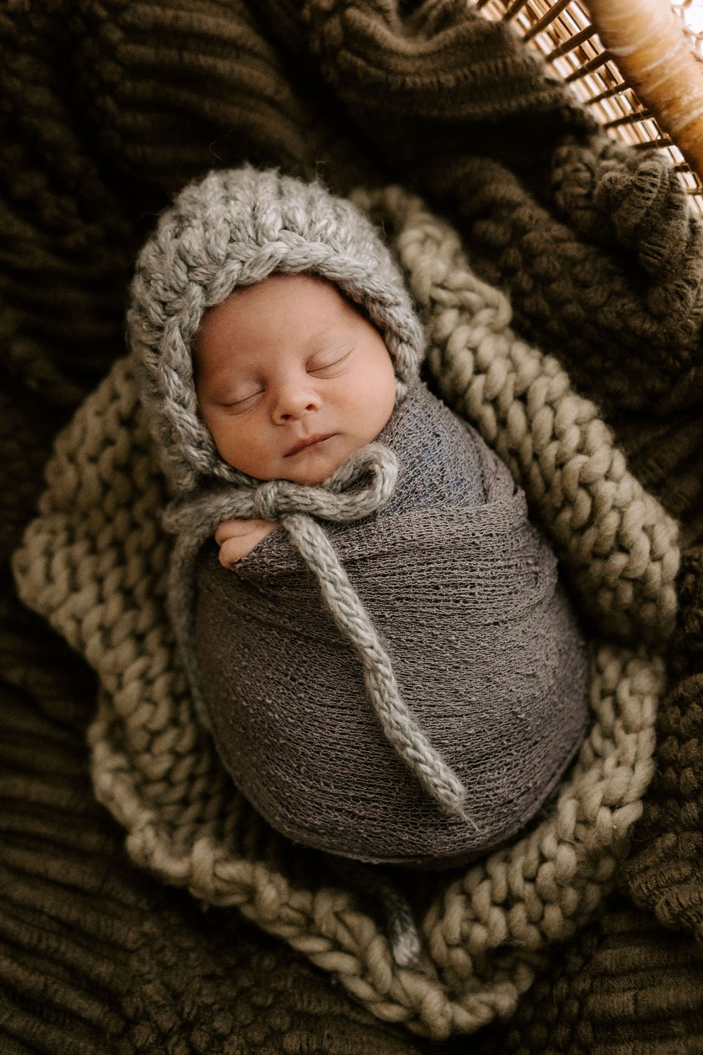 A sleeping newborn wrapped in a gray knit swaddle and bonnet rests on a chunky knit blanket.