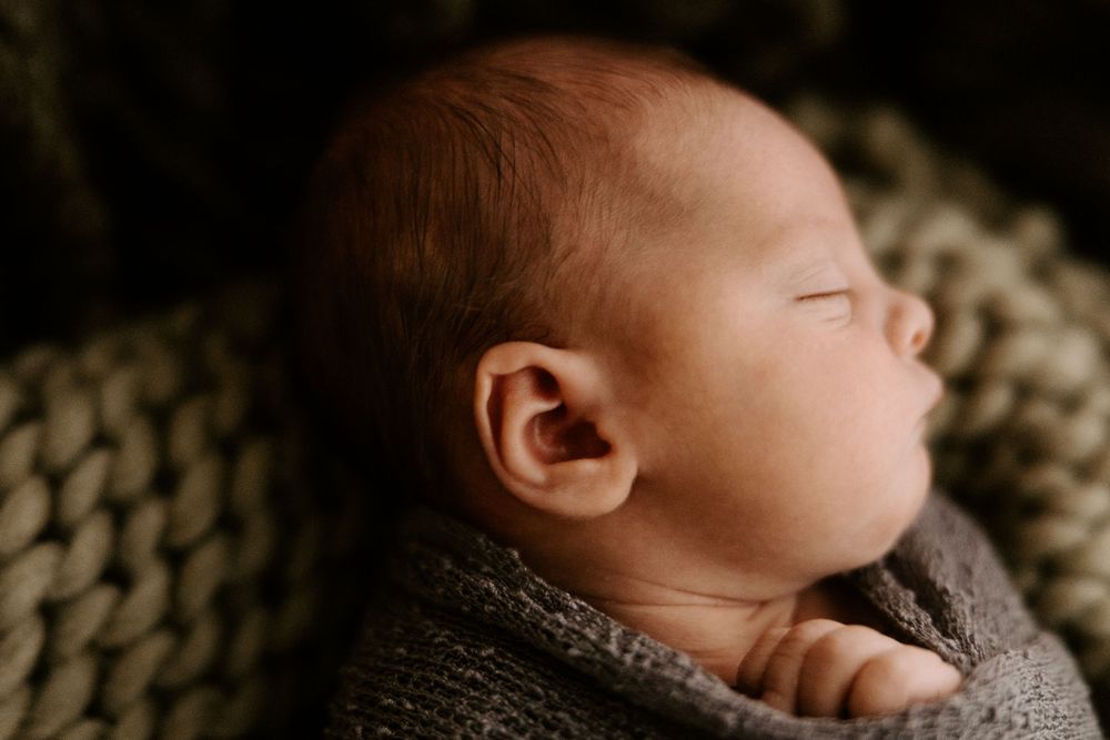 A newborn baby sleeps peacefully on a textured beige blanket in soft natural lighting.