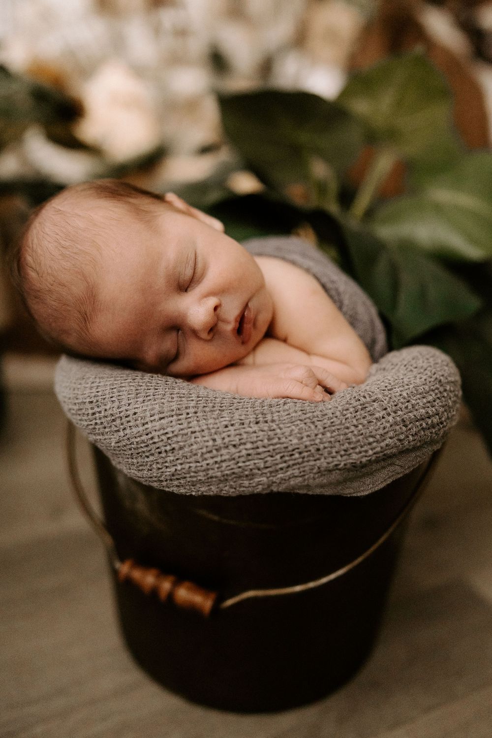 A sleeping newborn baby lies peacefully in a gray knit blanket inside a dark wooden basket.