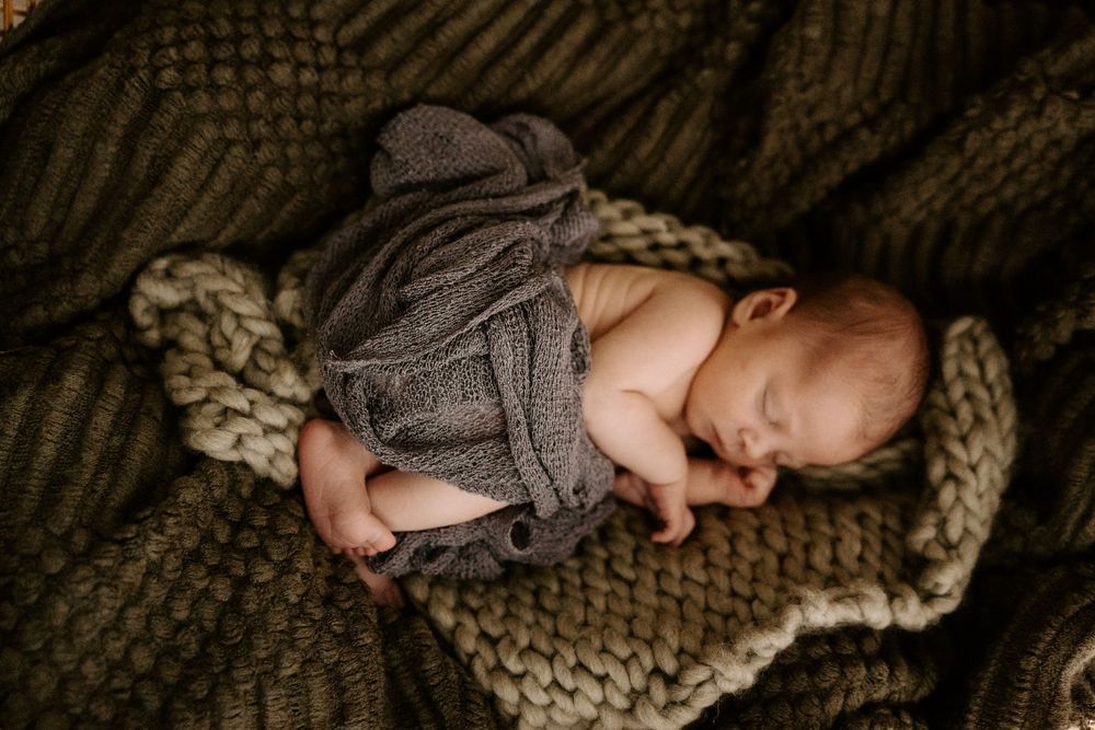 Newborn baby sleeping peacefully on chunky knit brown blanket while wrapped in soft gray swaddle.