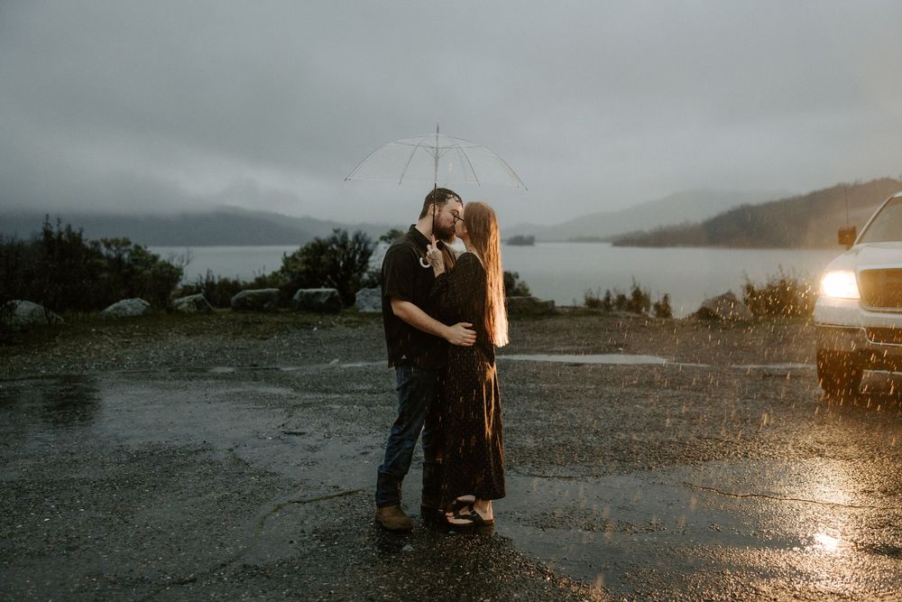 A couple embraces on a misty overlook at sunset with a dramatic mountain lake landscape in the background.