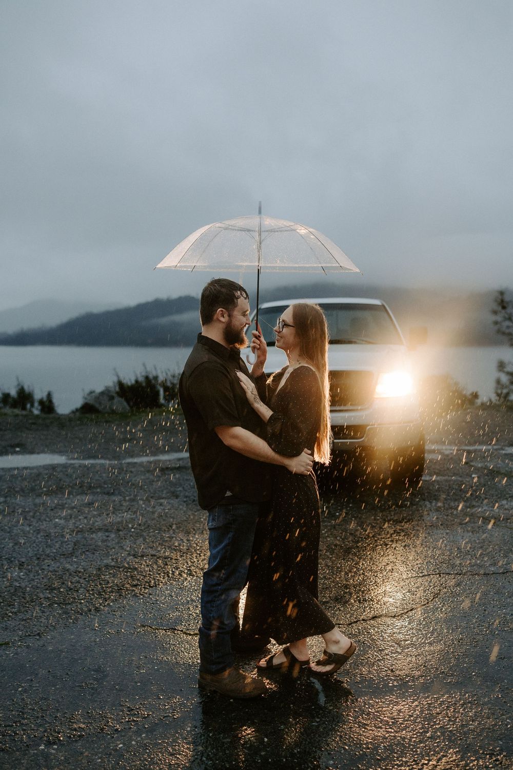 A romantic couple dances in the rain under an umbrella with car headlights illuminating the scene at dusk.