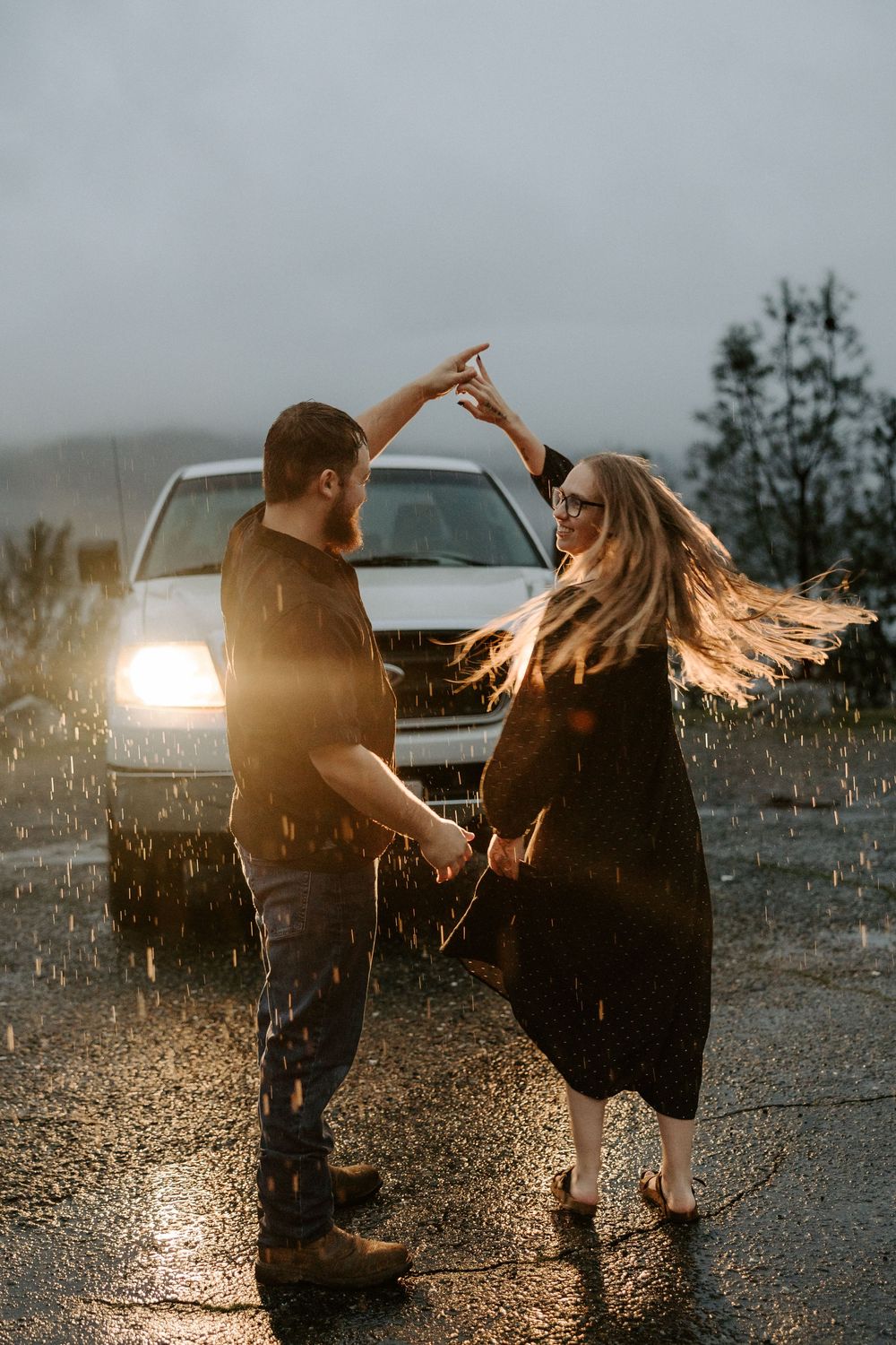 A couple twirls and dances playfully in the rain with dramatic car headlights creating a backlit silhouette.