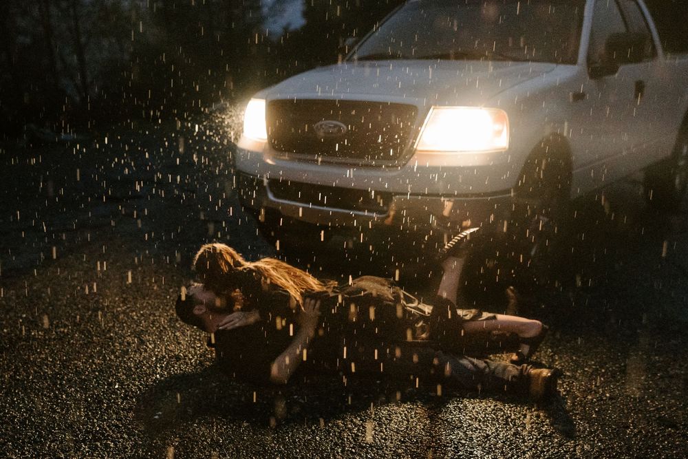 Raindrops splash on a car windshield at night while headlights illuminate the dark road ahead.