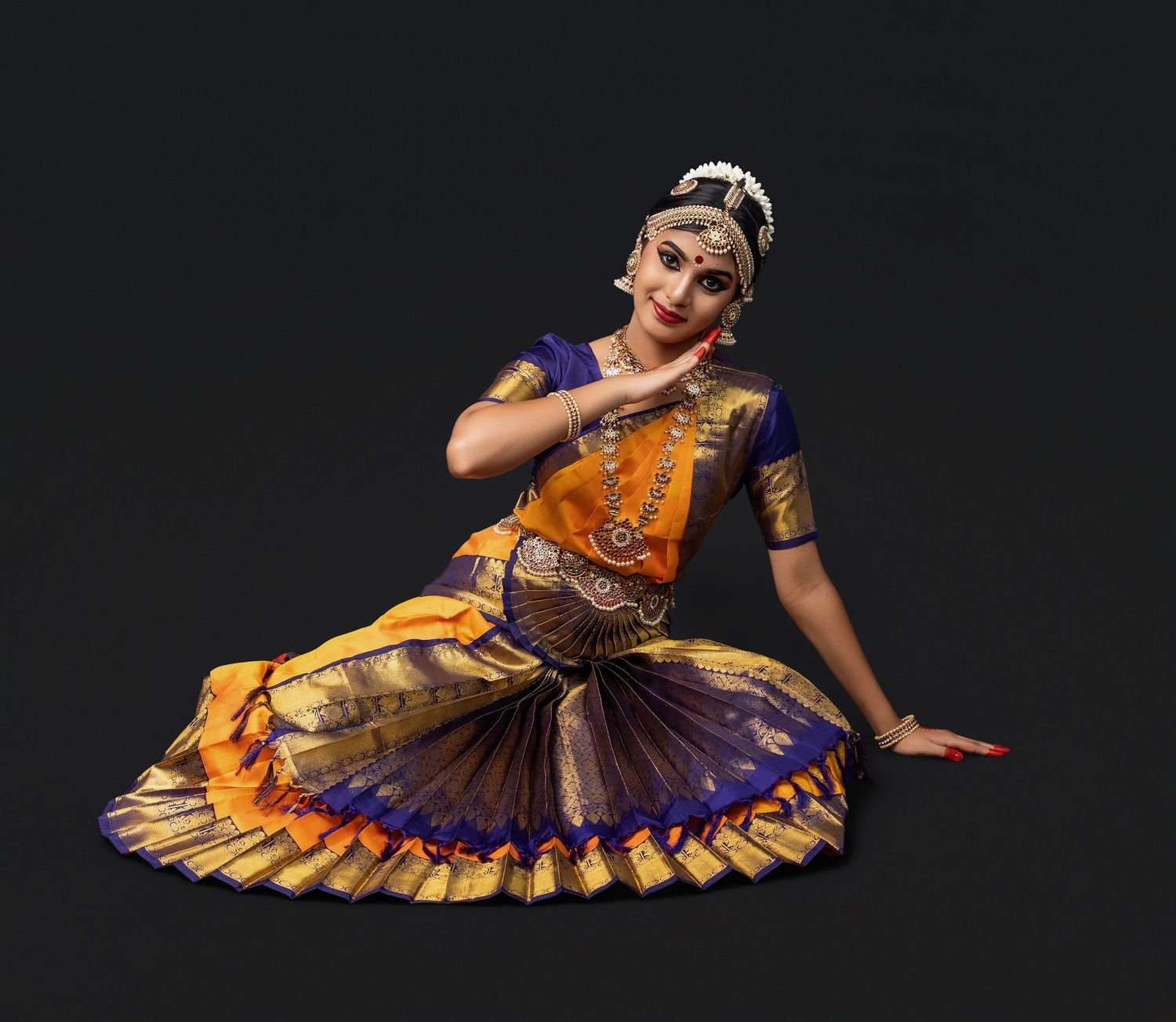 A classical Indian dancer in vibrant orange and purple traditional costume performs a graceful Bharatanatyam pose.