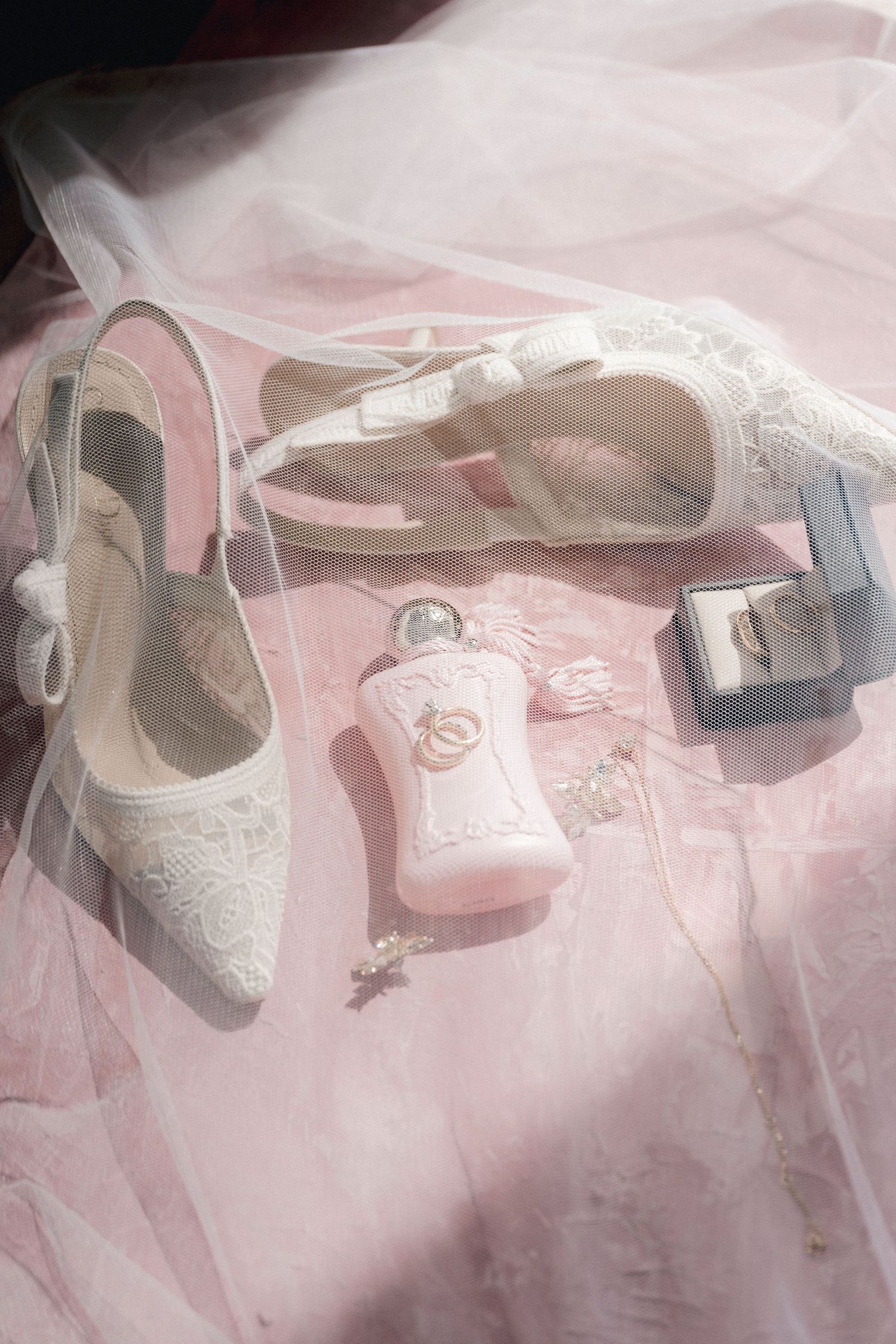 Elegant white lace bridal shoes and accessories arranged on soft pink tulle fabric create a romantic wedding styling scene.