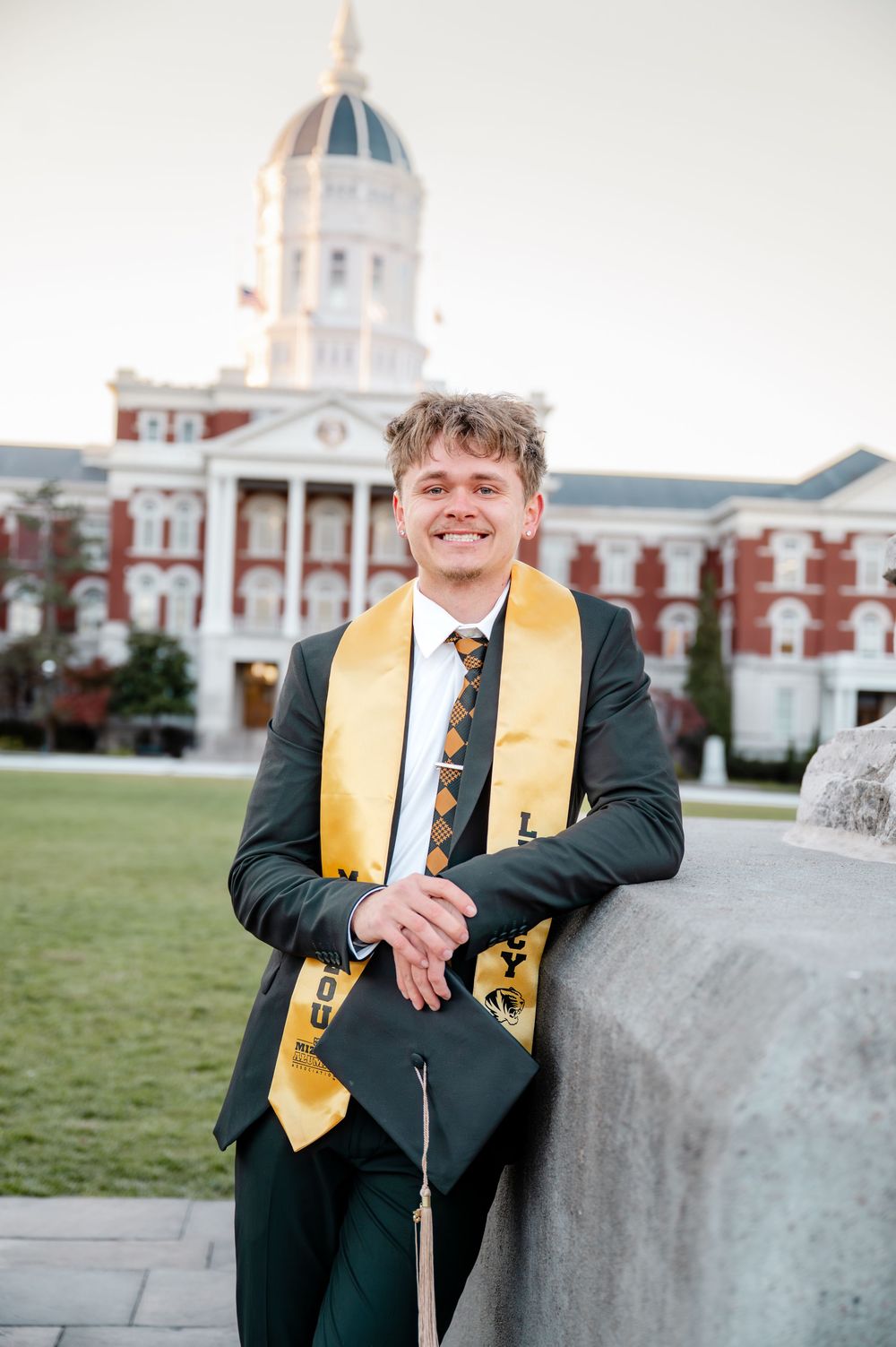 Graduate in cap and gown poses in front of historic university building with dome architecture.