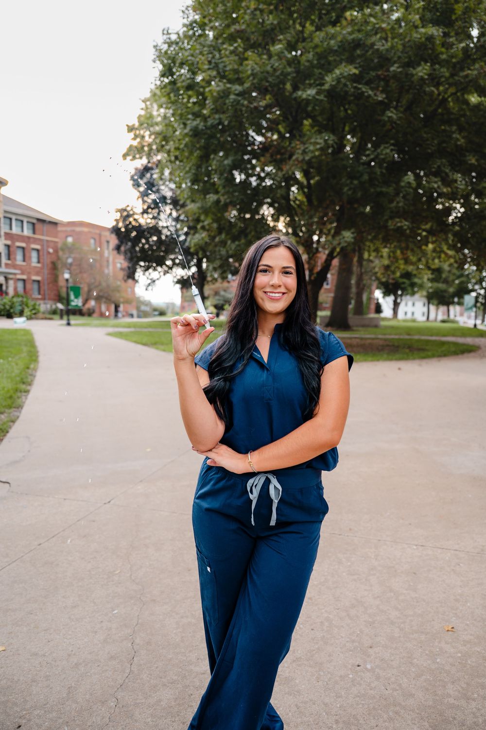 Healthcare worker in blue scrubs poses outdoors on university campus walkway.