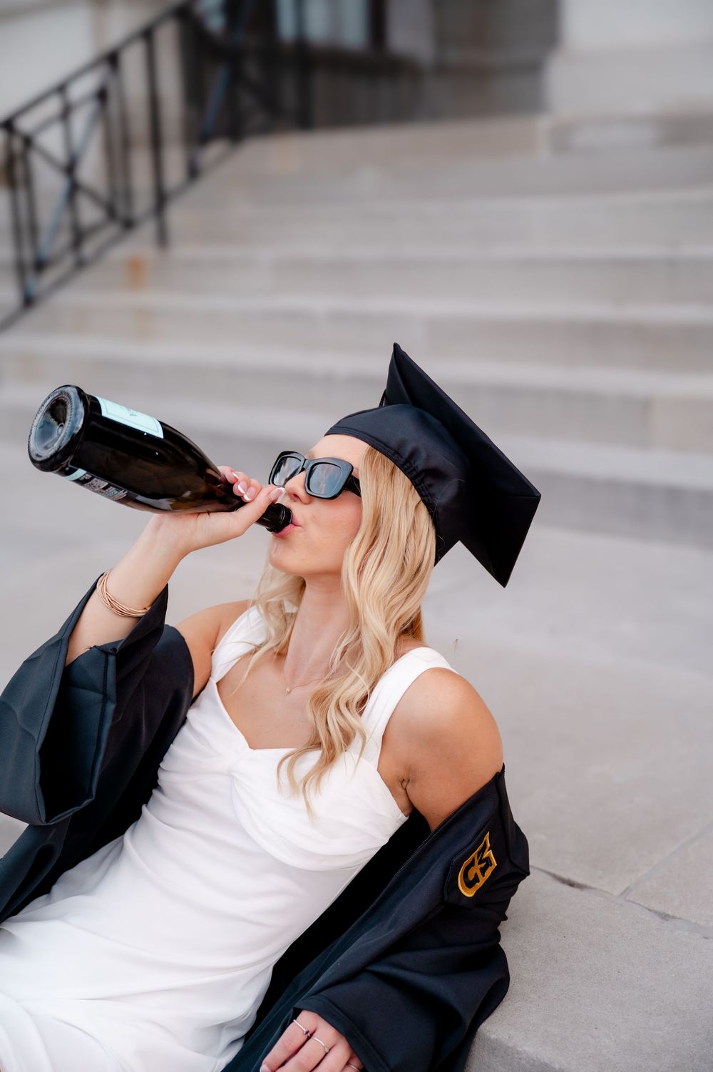 Graduate in cap and gown drinks from bottle while sitting on stone steps.