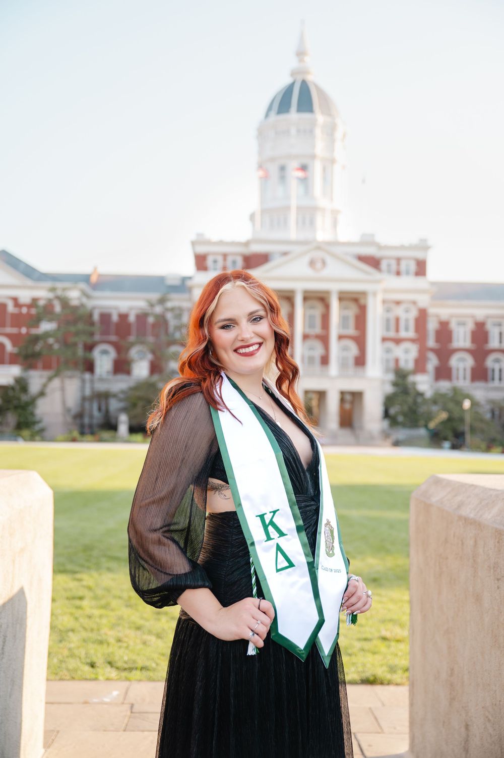 Graduate in academic regalia poses in front of historic university building with sorority sash.