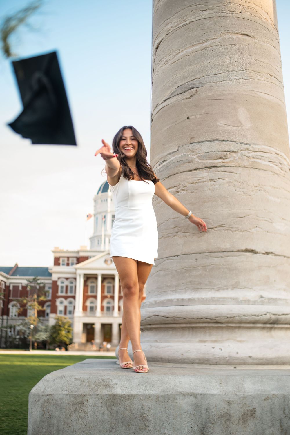 Graduate poses playfully by large stone column on university campus.
