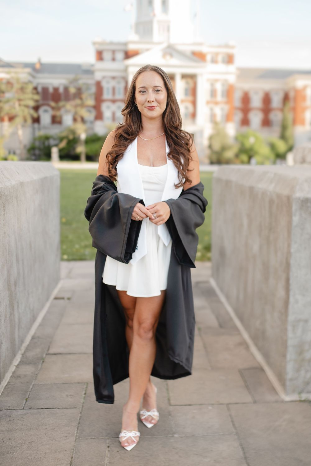 Graduate in formal attire poses on concrete walkway with building architecture in background.
