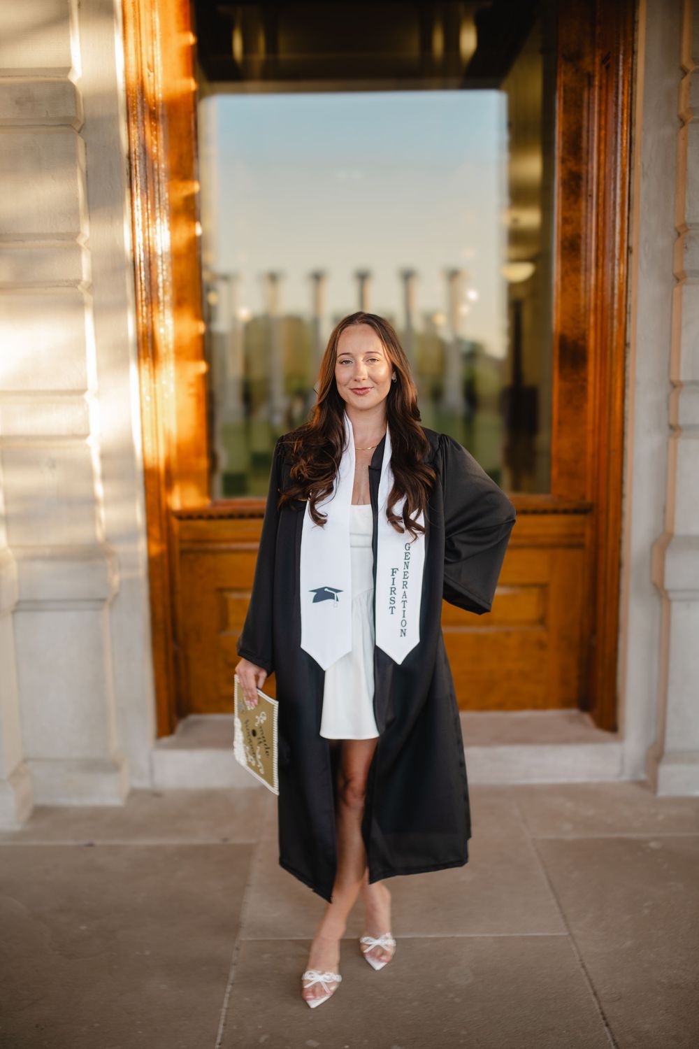 Graduate in black blazer and white dress poses in front of ornate building columns.