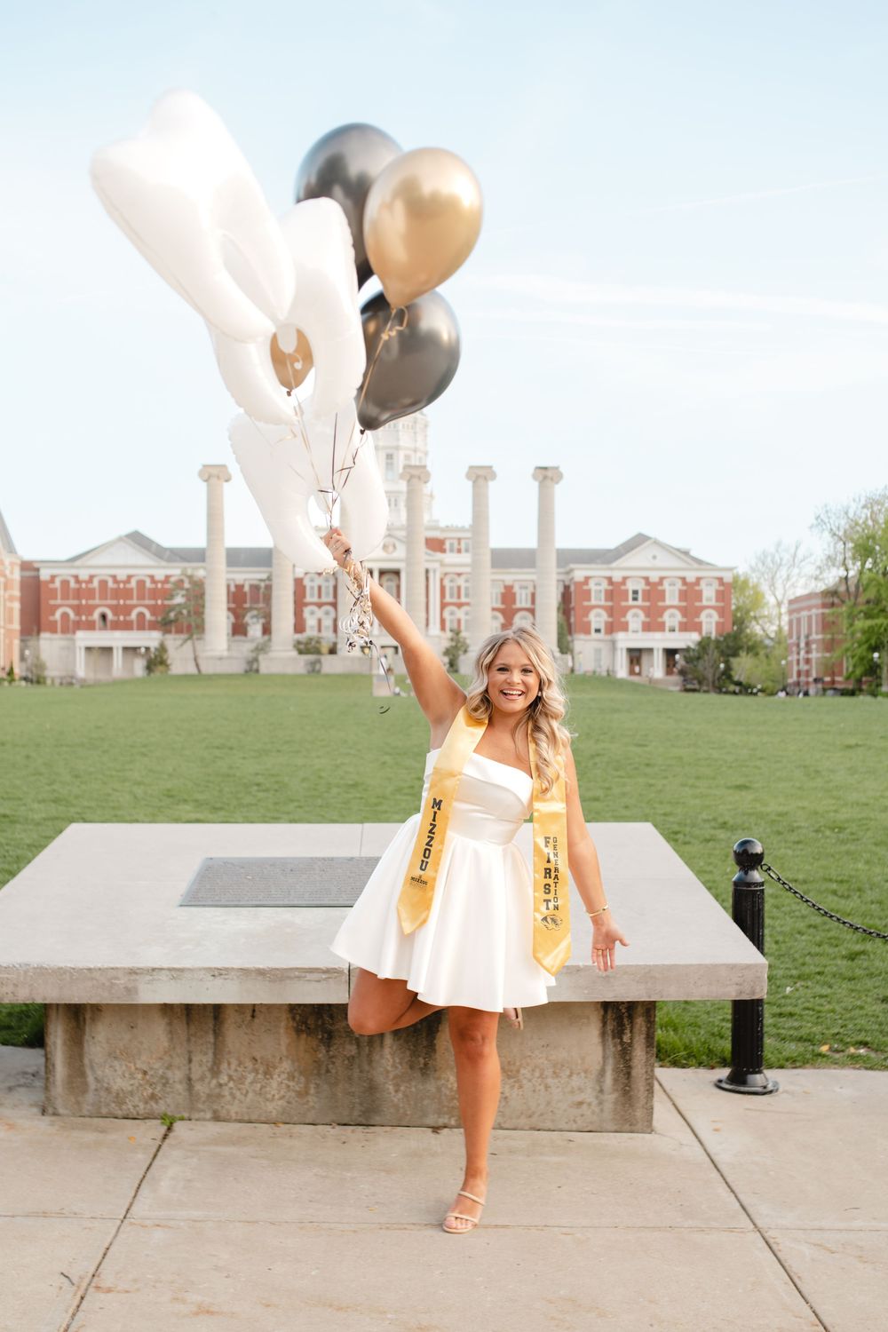Graduate celebrating with balloons on campus lawn wearing white dress and gold stole.