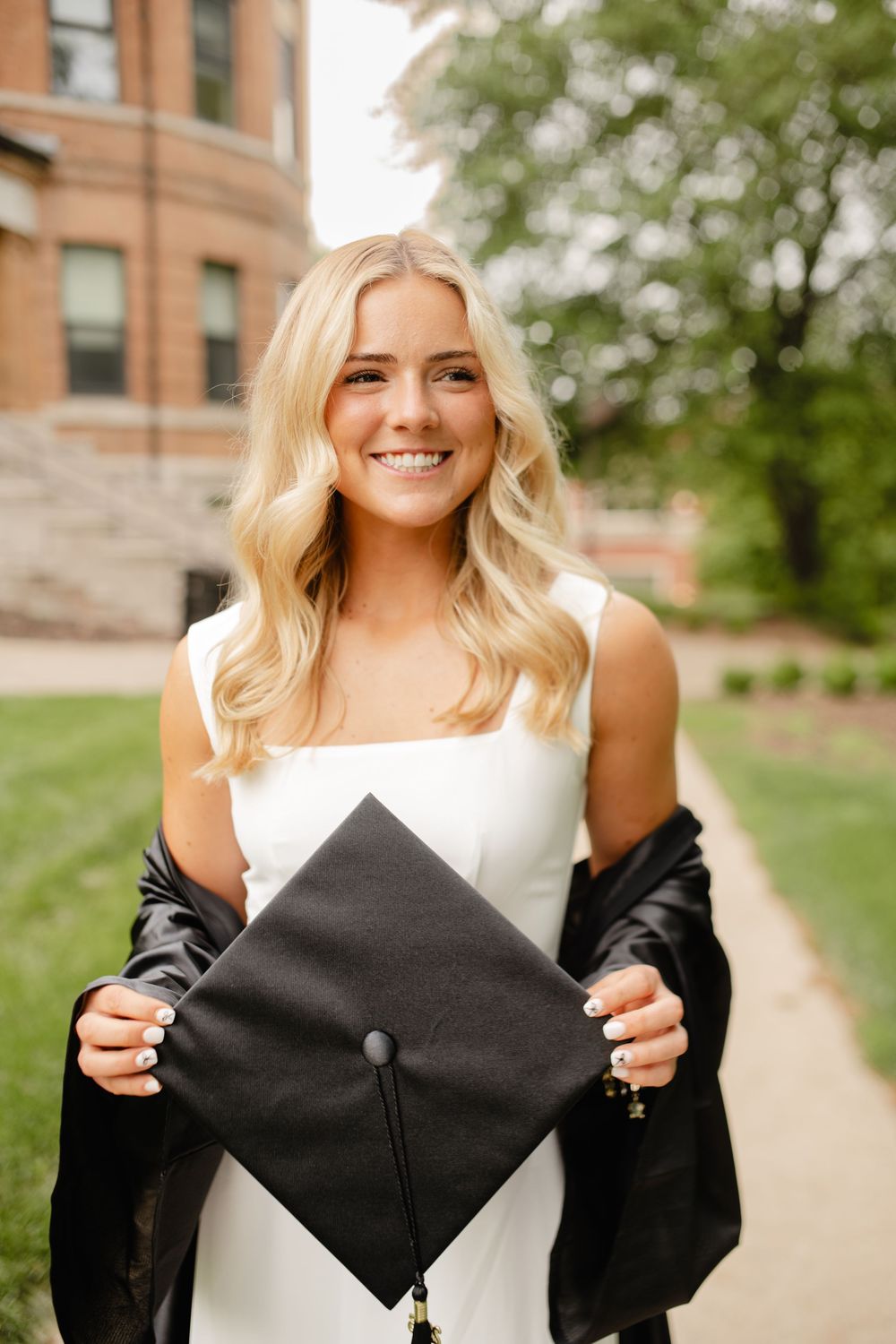 Graduate holding mortarboard cap wearing white dress and black graduation gown.