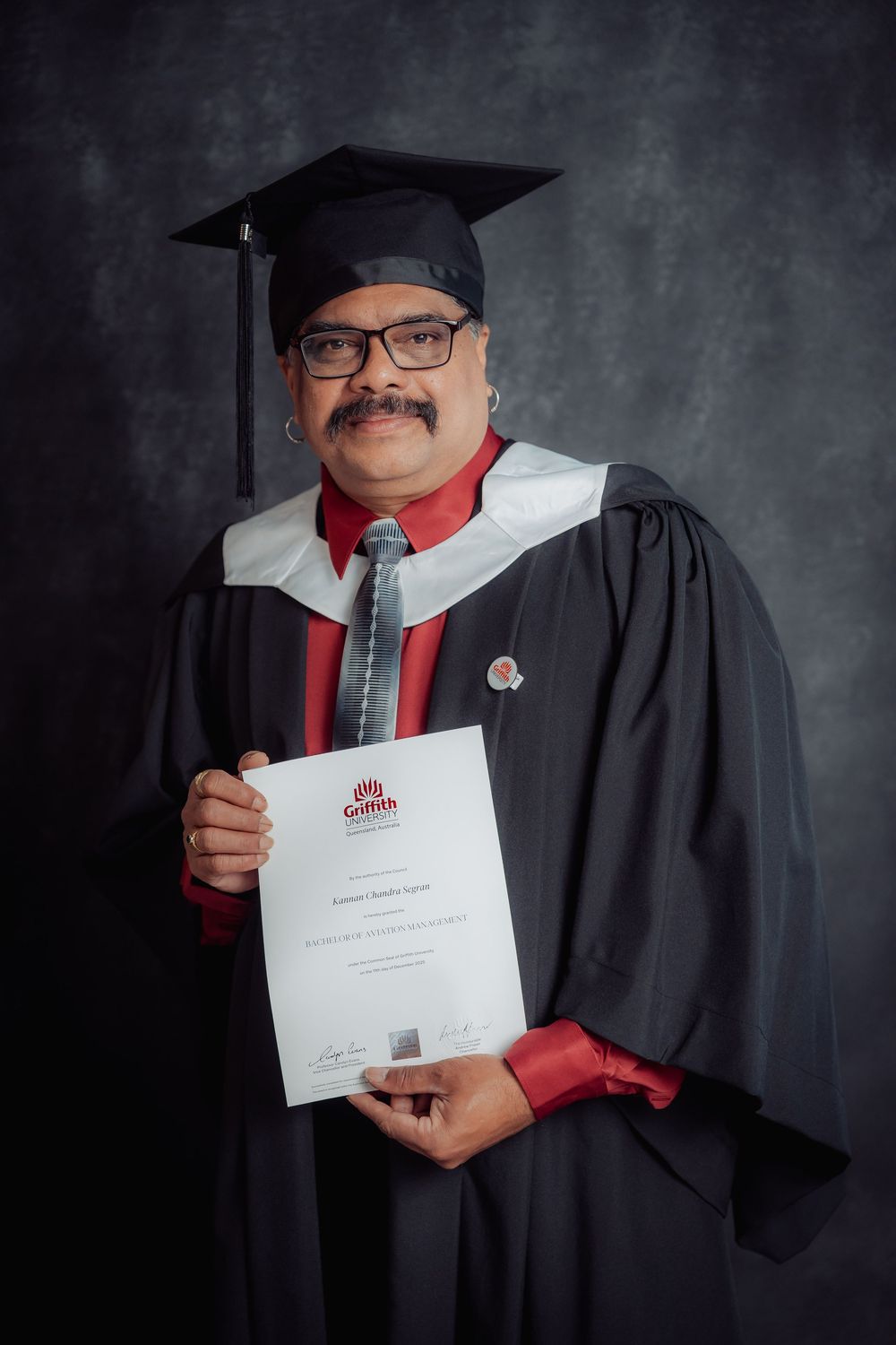 Graduate in black cap and gown holding diploma against dark background.