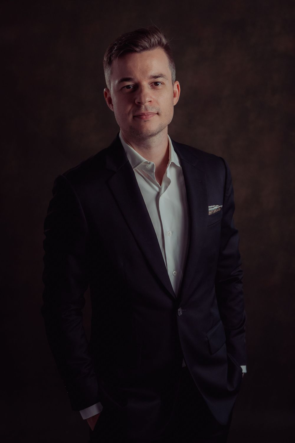 Professional headshot of person in dark suit and white shirt against moody dark background.