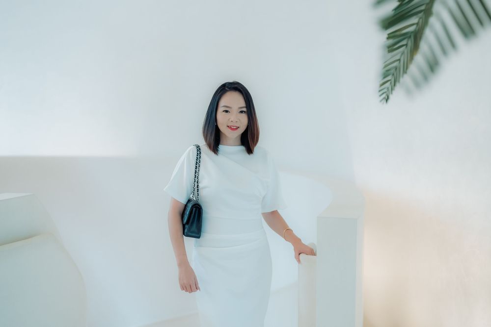 Someone in an elegant white dress and black handbag poses against a white wall with tropical palm leaves visible.