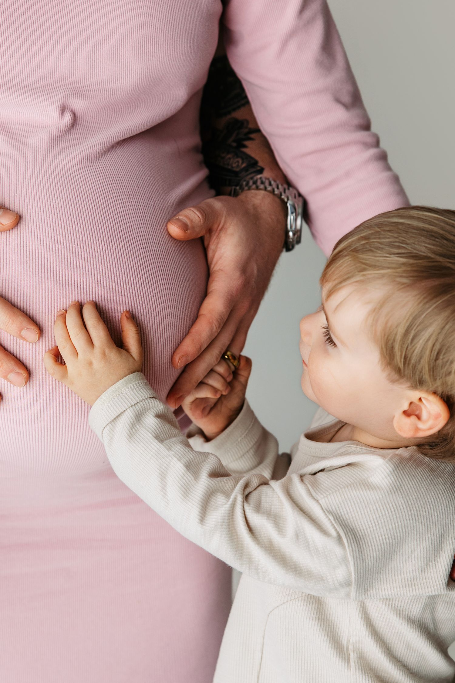 A young child in a white sweater touches a pregnant belly wearing a pink top while holding hands tenderly.