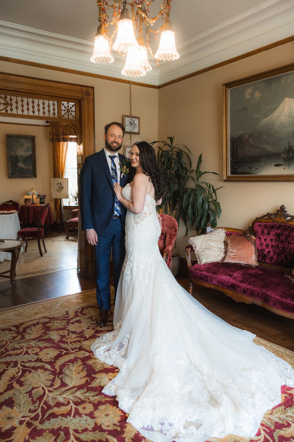 A wedding couple poses in an elegant Victorian-style room with ornate furniture and a sweeping white gown.