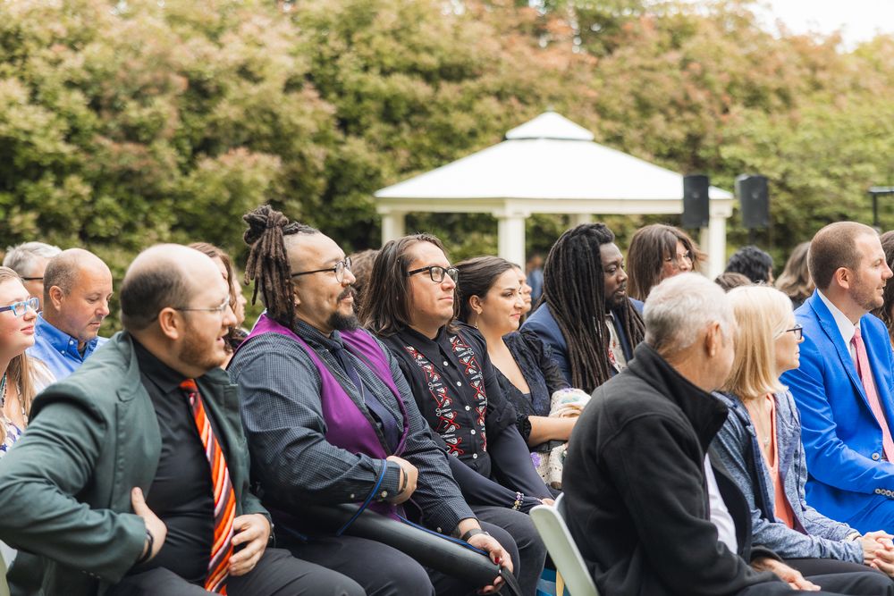 A diverse group of guests seated outdoors at a wedding ceremony under a white canopy on a sunny day.