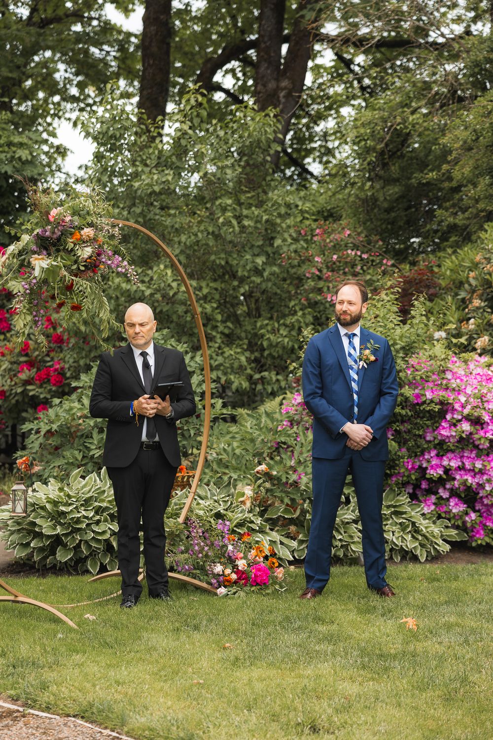 Groomsmen in blue suits stand casually on grass near flowering bushes and garden arch.