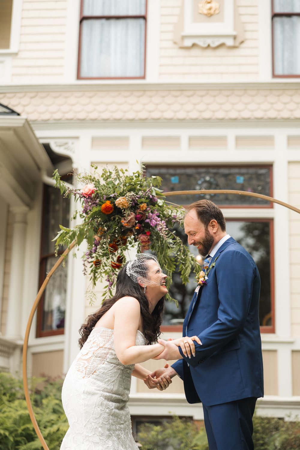 Wedding ceremony moment under a wooden arch decorated with burgundy and green florals against a white building.